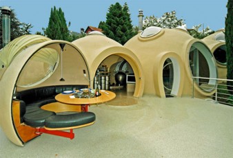 photo de la terrasse de la maison bulle de Hélène et Christian Roux à Fontaines-sur-Saône près de Lyon. Sur cette photo on voit la salle à manger ouvrante, ouverte, avec sa table ronde et sa banquette demi-circulaire. Les portes de la maison sont comme des arches romaines, les fenêtres et baies sont rondes.