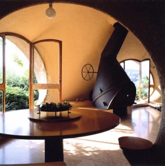 table ronde de 150 cm de diamètre réalisée en contreplaqué par Antonio Benincà avec son pied central réalisé avec un tube en métal, une fenêtre baie ronde avec une porte ouverte à gauche permet de sortir sur la terrasse, au fond la cheminée réalisée avec la récupération d'une queue de bétonnière de chantier inversée et sa porte réalisée avec un bras de suspension de Citroën 2 cv