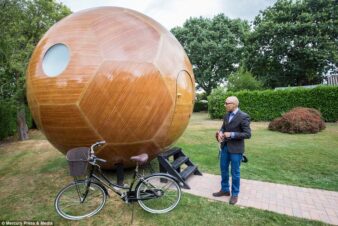 sur cette image, Jag Virdie le créateur du Conker living, contemple une sphère en bois vernis de ses créations, d'un diamètre d'environ 4 mètres. La sphère est posée dans un jardin, avec la bicyclette de Jag à côté. Jag Virdie en jean's et veste personnalisée conker living