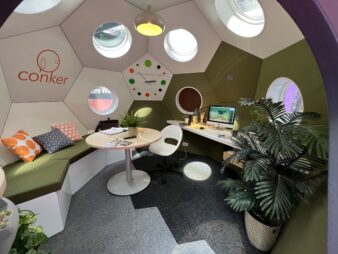 interieur d'une mini maison conker living de Jag Virdie, un espace bureau vivant, avec le bureau à droite où l'on voit un imac apple et accessoires de travail, des plantes vertes, des murs couleurs vert kaki et crème, le logo conker living sur une alvéole, les hublots qui réfléchissent la lumière au sol, un coin salon avec coussins en demi cercle couleur kaki et coussins colorés orange