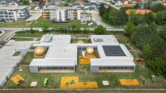 cette photo présente une vue aérienne de la crèche de Székesfehérvár en Hongrie, on y voit un batiment en béton entouré d'un jardin et de jeux pour enfants, entre la forêt et la ville, avec 3 sphères de couleur or surmontées d'un skydome vitré. Les bâtiments en béton accueillent 56 enfants. Il y a 2 préaux. Toute la crèche est entourée par un jardin.