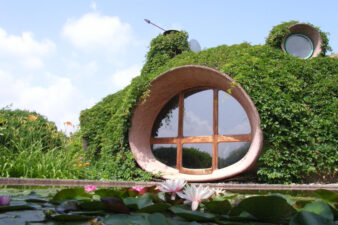 maison bulle antonio beninca, recouverte de végétation, avec son grand hublot facade sud, et son bassin à poissons rouges devant et ses nénuphars, le grand hublot en béton avec porte en bois contre plaqué, la girouette en haut sur le toit et le petit hublot de la chambre d'antonio en haut