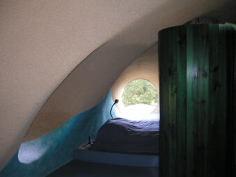 photo de la chambre d'Antonio Benincā à sa maison bulle de Saint Germain Laval dans la Loire. Au premier plan à droite, le retour du meuble dressing en bois peint en vert, a gauche on distingue de la lumière sortant du hublot, au fond un hublot qui éclaire le lit,tout ça sous une voute