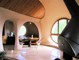 photo de l'intérieur de la maison bulle d'Antonio Benincà a Saint Germain Laval dans la Loire. On y voit de droite à gauche, d'abord sa cheminée fabriquée à partir d'une queue de bétonnière de chantier en inversée, la porte d'entrée ronde réalisée en contreplaqué par Antonio, puis la table ronde pour le repas avec un seul pied central et la banquette autour, et une claustra au plafond