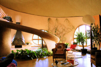 photo de l'intérieur de la maison bulle de Antonio Benincà dans la Loire. De gauche à droite, on aperçoit en fond la cheminée qu'il a fabriqué à partir d'une queue de bétonnière de chantier inversée, puis 2 portes ronde en bois, des plantes vertes, une lampe pétale sur pied création d'Antonio, des fauteuils club, et bien sur au centre de la pièce, le fameux tobogan qui sert d'escalier pour monter au à l'étage mais qui n'est pas un escalier, c'est un tobogan en voile de béton