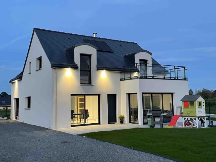 photo d'une maison traditionnelle, réalisée en parpaings, avec sa toiture en ardoises, deux fenêtres chiens assis dont une avec petite terrasse au dessus d'une veranda, le garage est derrière avec accès sur le côté, belle pelouse devant avec barbecue au gaz mobile et jeu pour enfants