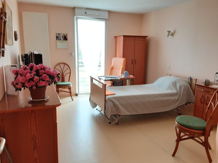 photo d'une chambre senior simple dans une maison de retraite, vide, avec un lit médicalisé vide, une armoire ikea fade, deux chaises, dans une ambiance morne