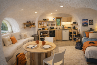 image de l'intérieur d'un studio bulle, créé par l'ia, avec le salon à gauche, la table ronde pour le repas devant et ses chaises, la cuisine au fond, et le coin pour dormir à droite, avec des murs et un plafond courbe et une fenêtre hublot à gauche