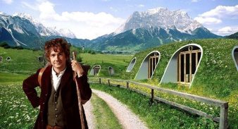 Bilbo le Hobbit devant des maisons Green Magic Homes semi-enterrées et recouvertes de végétation dans un paysage montagneux.